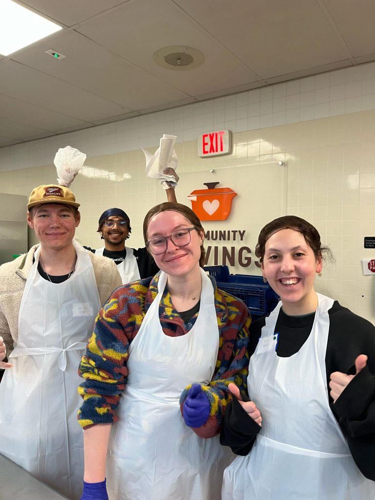 Three members of Valpo's Sweetwine Music Ministry posing for a photo in hair nets and caps in a kitchen where the group is labeling food items, with Fox Wilmot '24 smiling in the background.