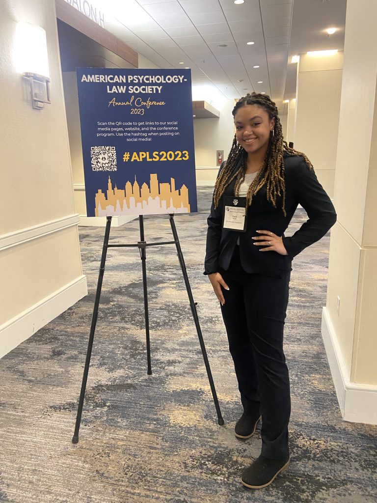 Kayla Smith '24 smiling and posing next to a sign that reads "AMERICAN PYSCHOLOGY-LAW SOCIETY ANNUAL CONFERENCE 2023."