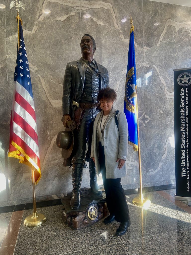 Kayla Smith '24 smiling in front of a larger-than-life bronze sculpture, titled "Frontier Marshal" inside the United States Marshals Service headquarters in Crystal City, Virginia.