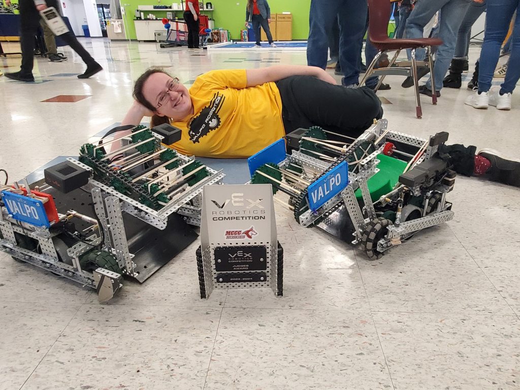Lauren Kadlec '24 posing on the floor of the campus building beside two of the Valpo Robotics robots and an award plaque from the Vex Robotics Competition.