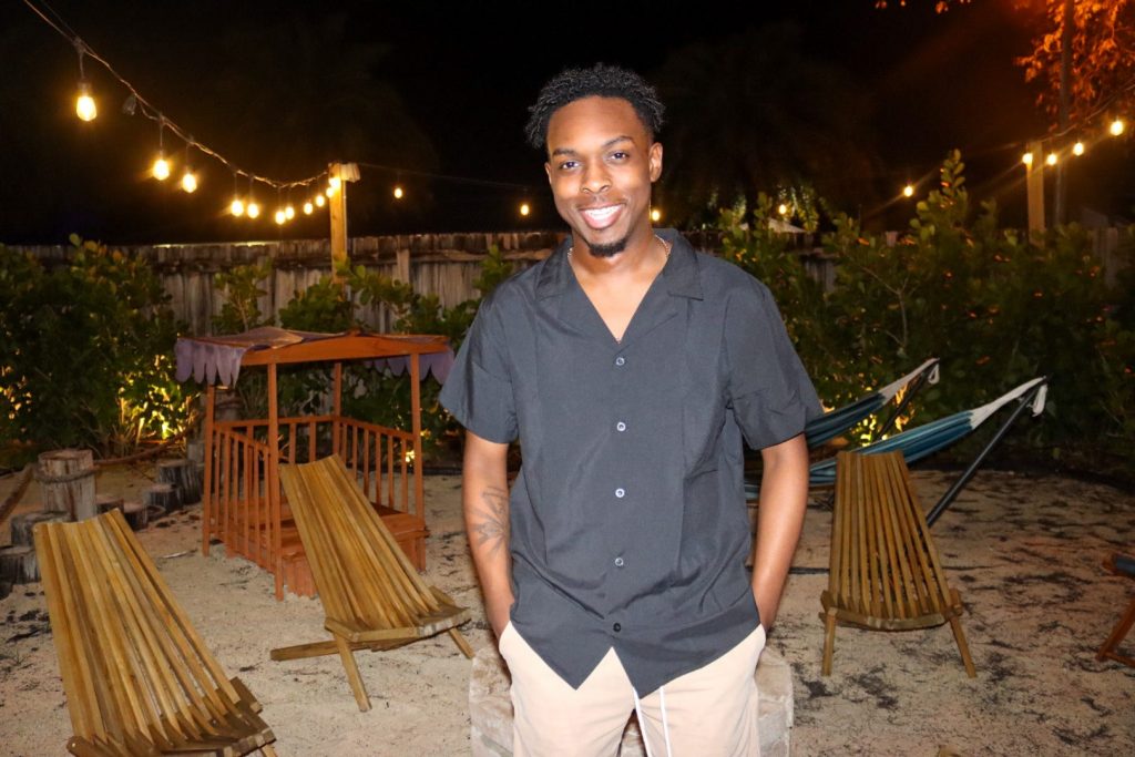 Valpo graduate Jaylen Jude '24 smiling for the camera with his hands in his pockets on the beach at night, with greenery, hammocks, lounge chairs, and string lights in the background.