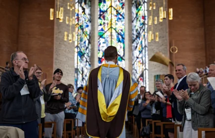 A person in academic regalia stands in a church as people around them clap and stained glass shines behind, celebrating the Inauguration of President Rev. Brian Konkol at Valparaiso University.