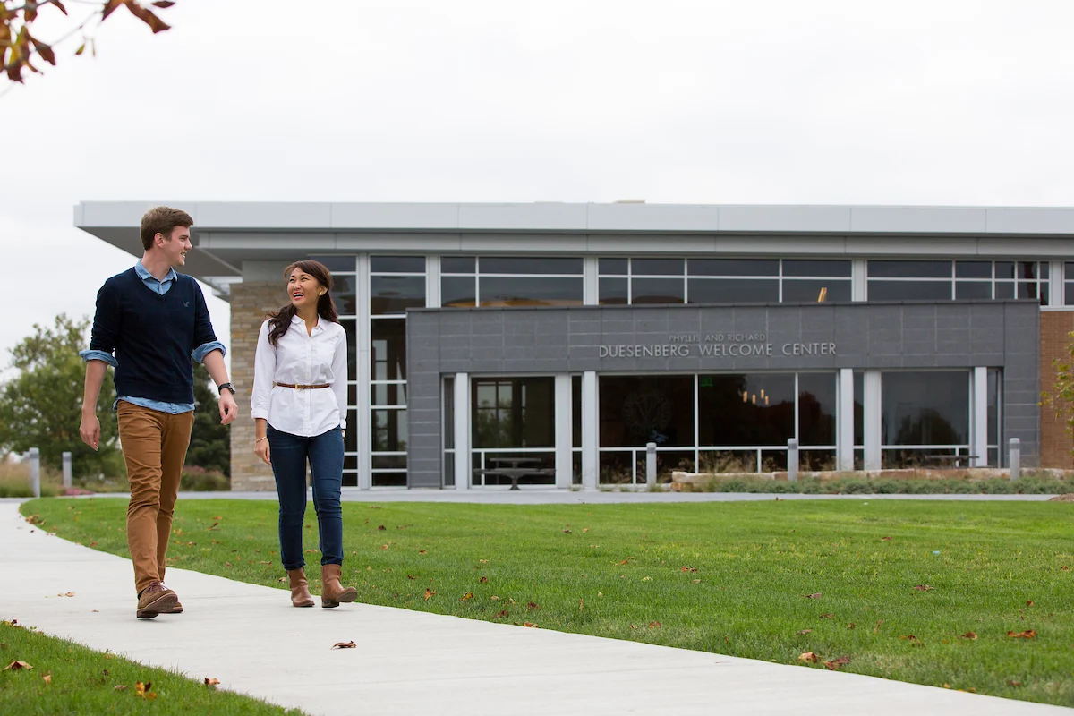 Two people walk on a sidewalk in front of a modern building labeled Duesenberg Welcome Center.