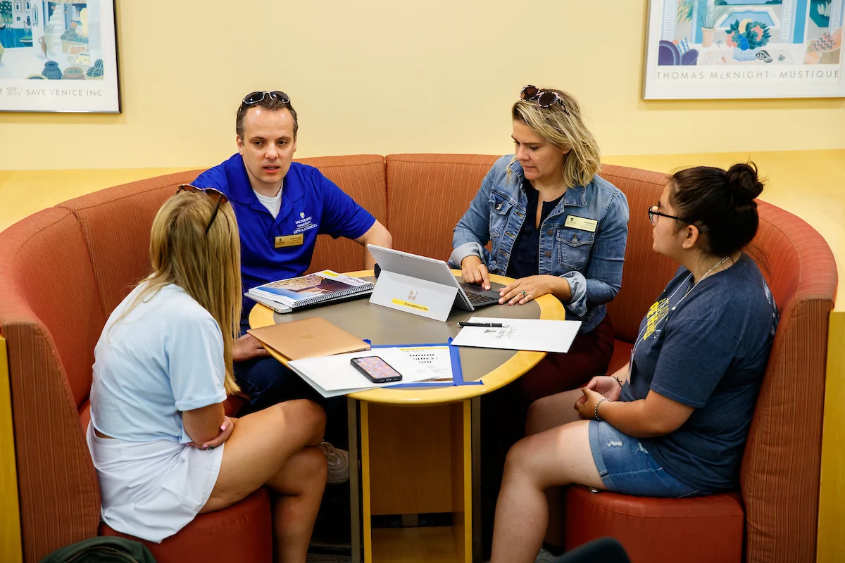 Four people sit in a curved booth having a discussion with laptops and papers on the table.