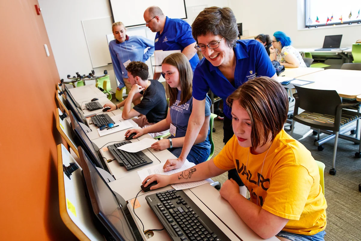 Students work at computers in a classroom while instructors assist and observe, everyone focused and engaged.