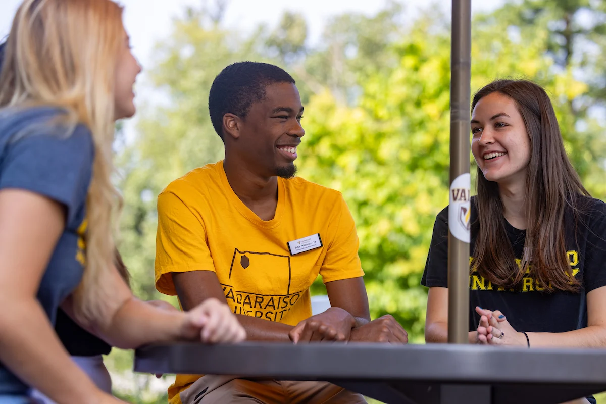 Three students wearing university shirts sit at an outdoor table, smiling and talking together.