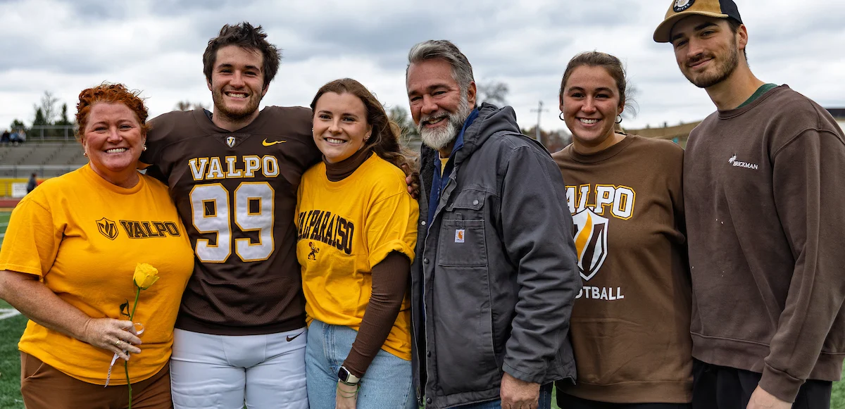 Six people standing together on a field, smiling, wearing Valpo apparel, one holding a yellow flower.