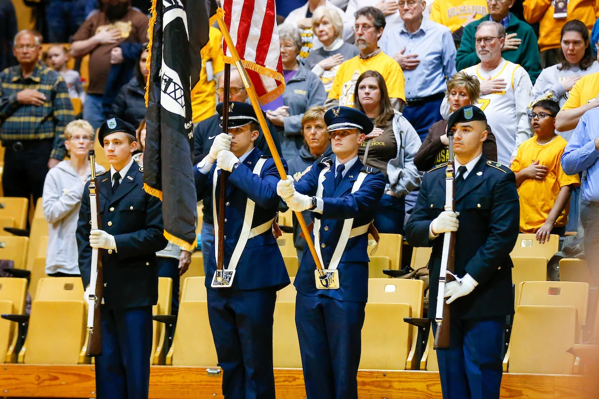 Four uniformed honor guards hold flags during a ceremony, as people stand with hands over their hearts.