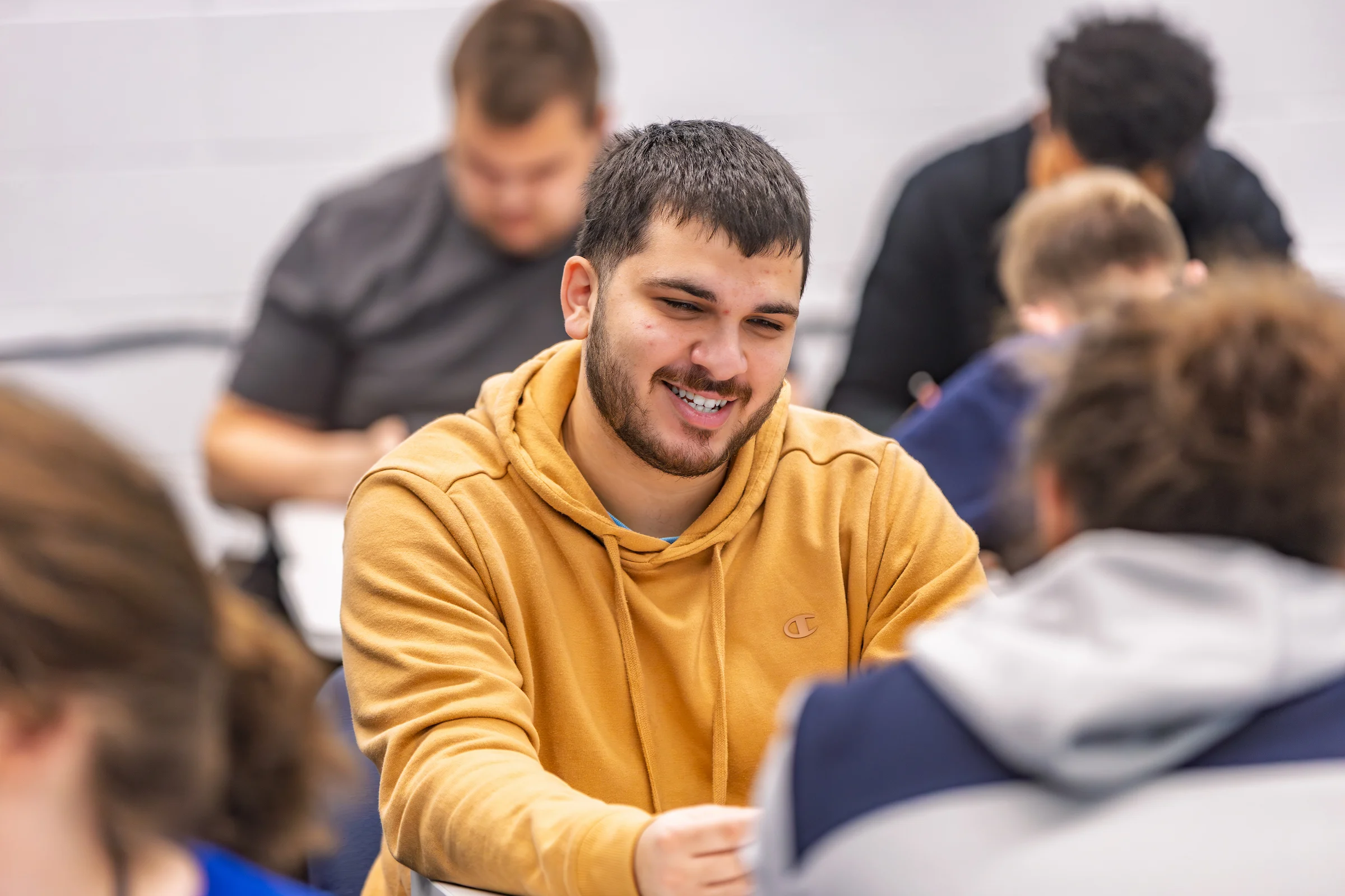 Man in a tan hoodie smiles and talks with others in a classroom setting.