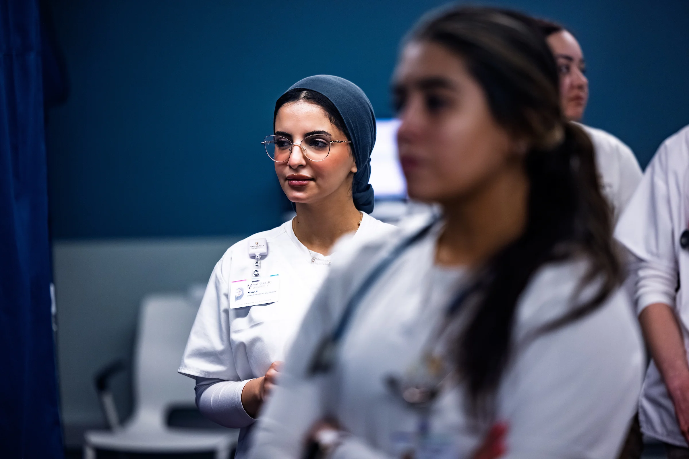 A group of nurses in white uniforms listens attentively in a hospital room.