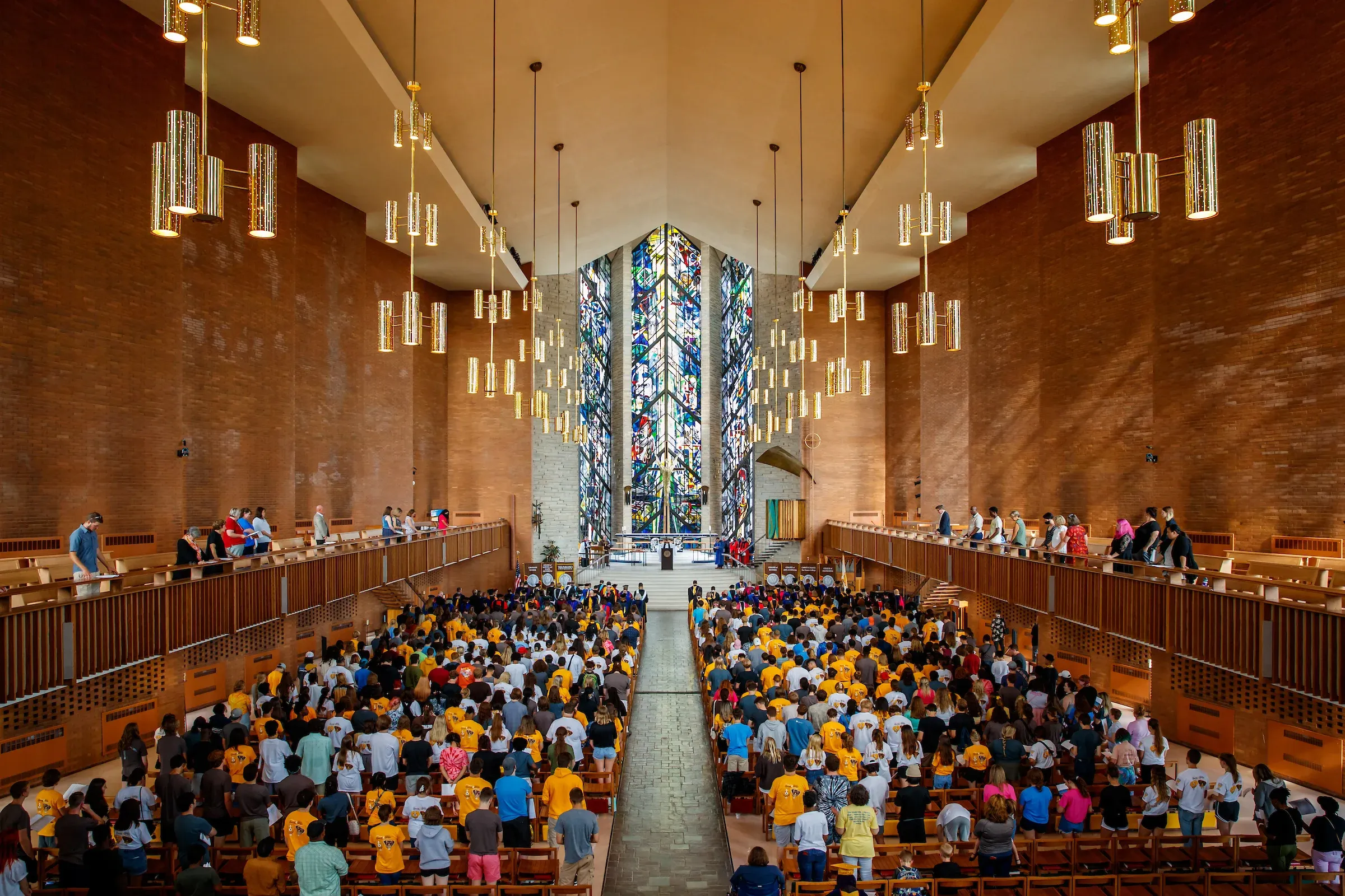 Large crowd in a church with stained glass windows and hanging lights, viewed from the back.