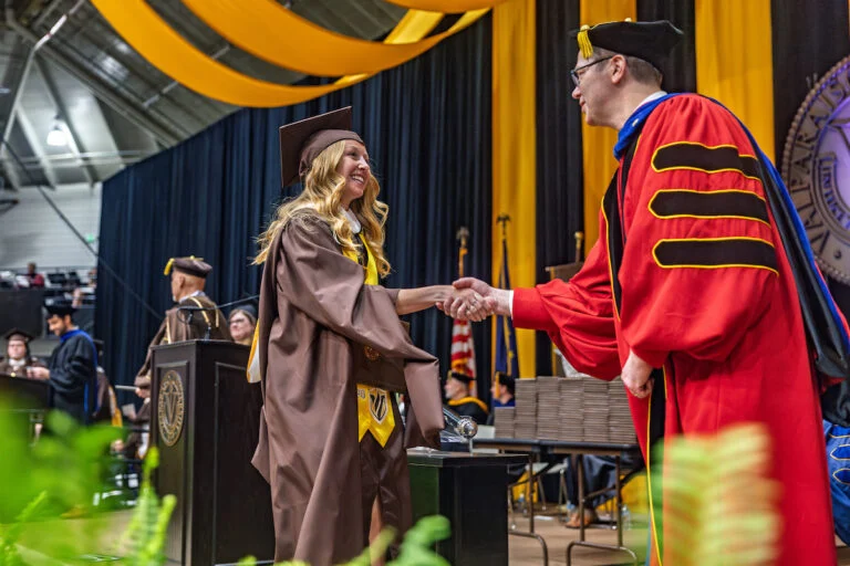 Graduate in brown cap and gown shakes hands with faculty member in red robe at a graduation ceremony.