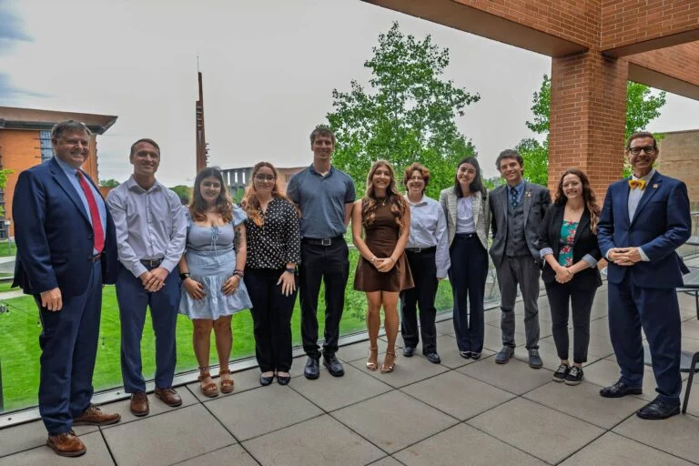 A group of eleven people pose together on a terrace with buildings and trees visible in the background.