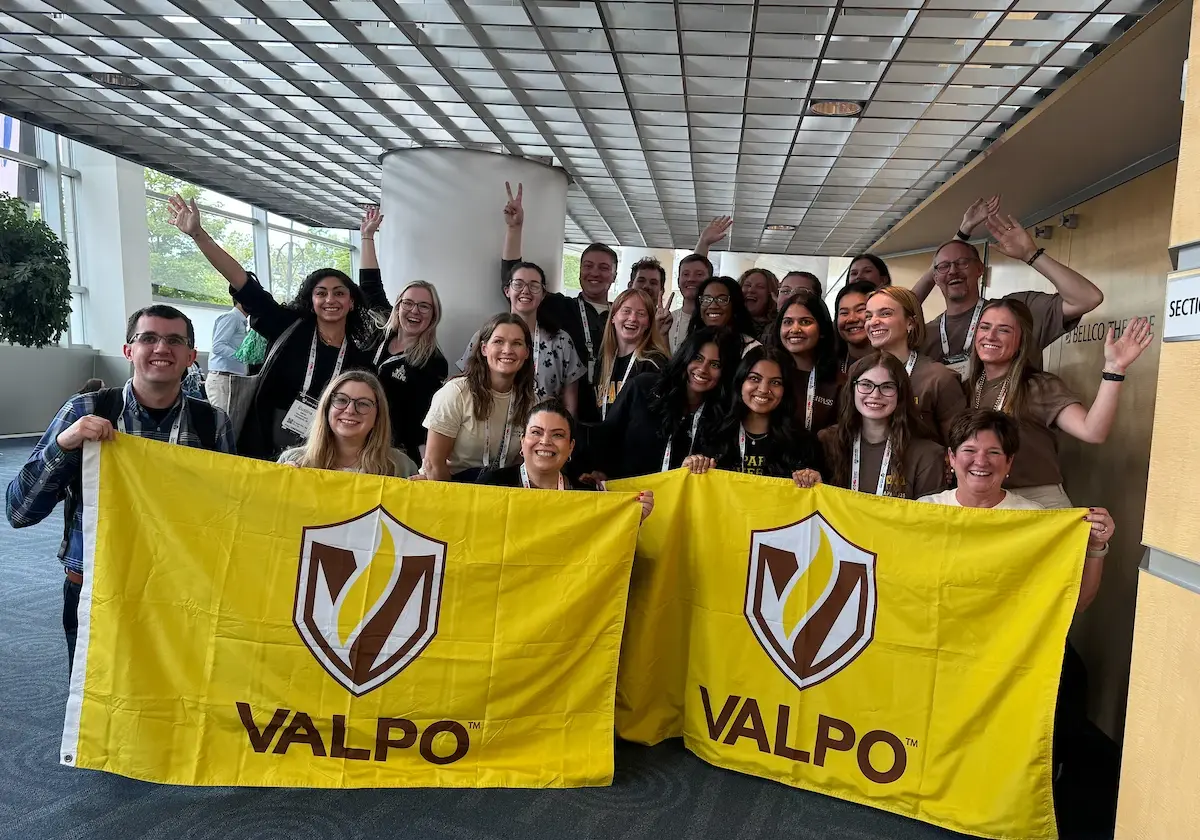 Group of people smiling indoors, holding two yellow “VALPO” flags with a shield logo, waving at the camera.