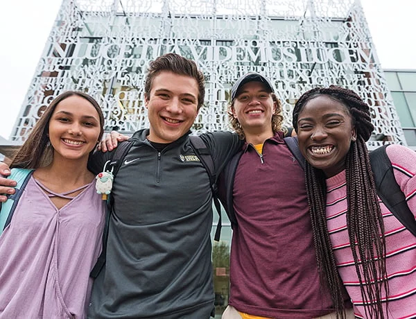Four smiling students stand arm-in-arm outside a modern building with large lettering on its facade.