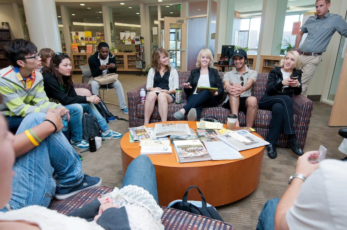 A group of international students sits in a circle talking in a library or lounge, with papers spread out on a table in the center.