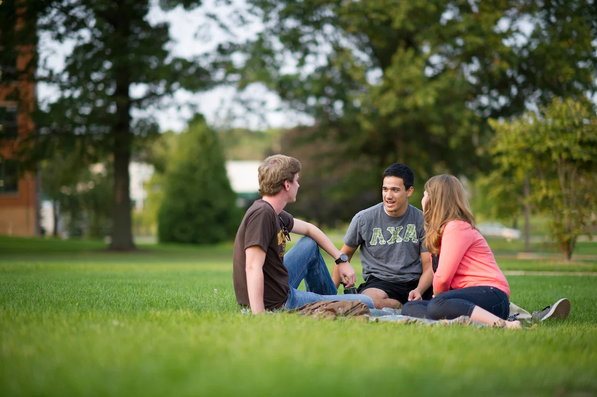 Three people sit on grass in a park, talking and smiling on a blanket, trees in the background.