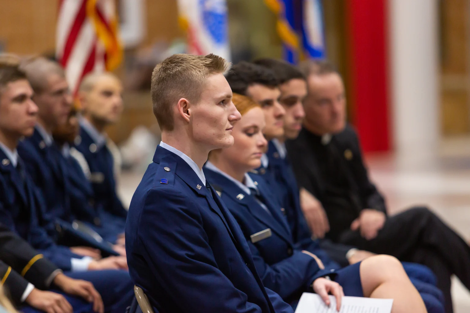 Group of people in formal military uniforms sit in rows, facing forward at an indoor event or ceremony.