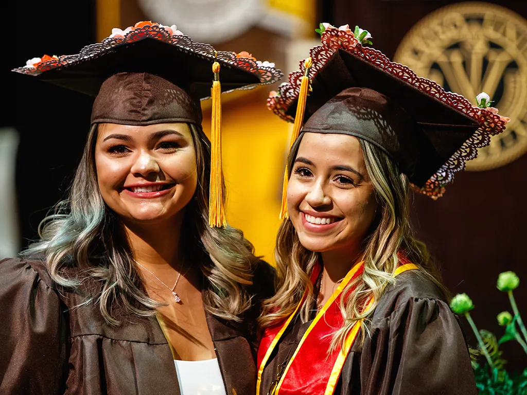 Two smiling graduates in caps and gowns pose together at a graduation ceremony.