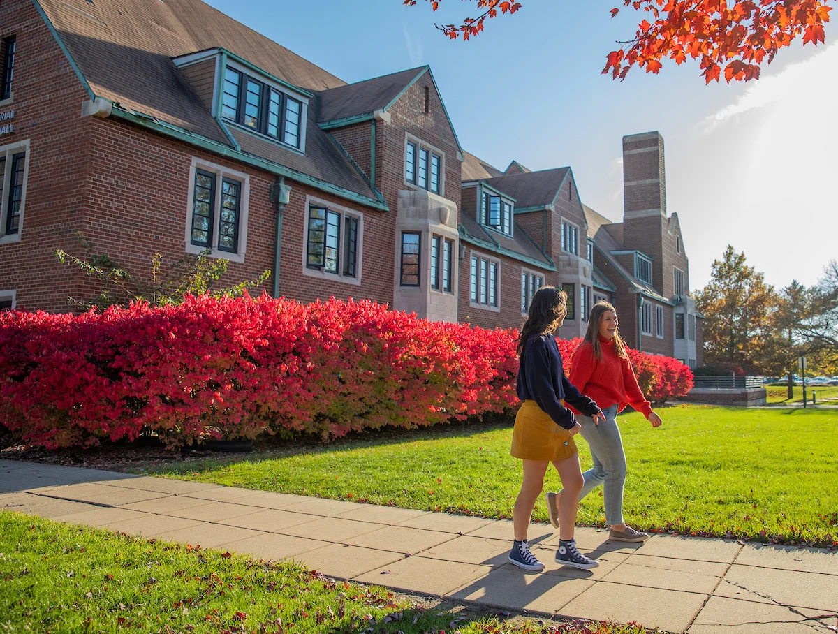 Two people walk on a sunny sidewalk by a brick building, red shrubs, and autumn trees.