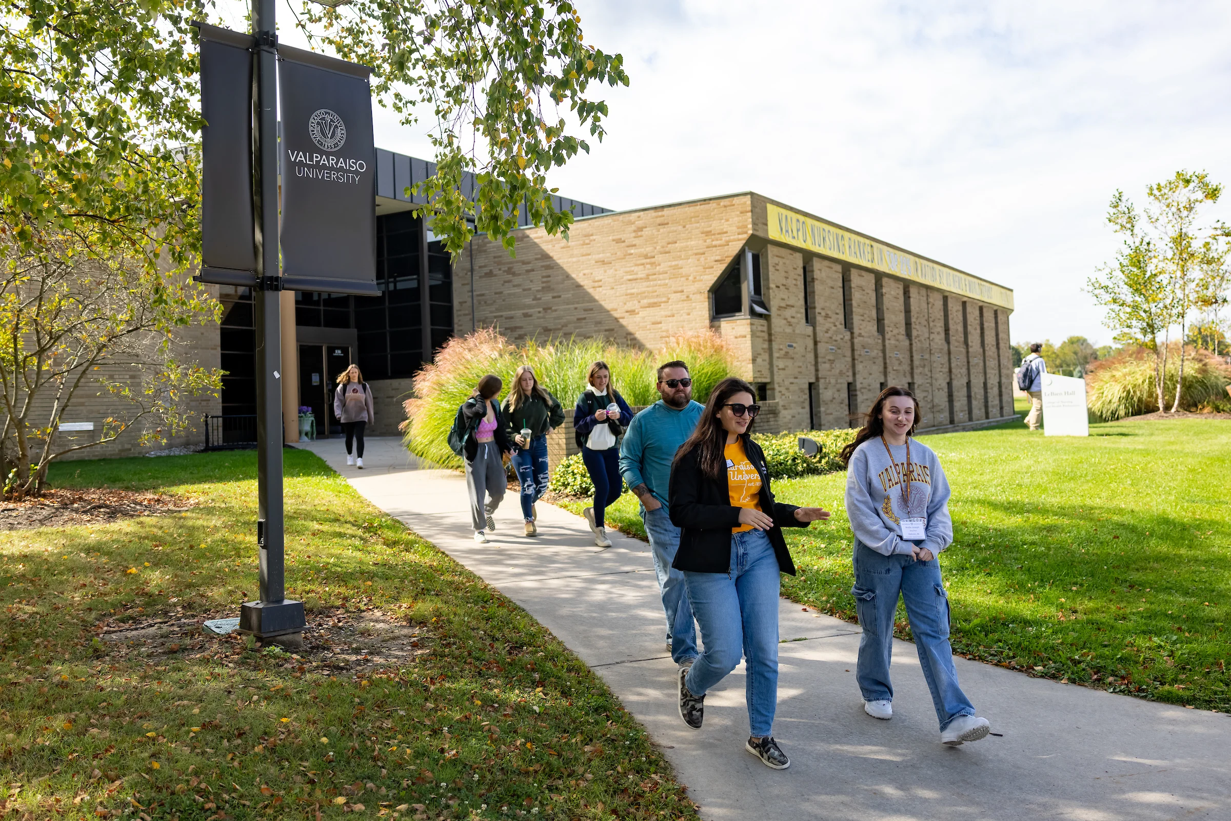 Students walk on a sidewalk outside Valparaiso University on a sunny day, passing a campus building as conversations about the Financial Aid Application and Financial Aid Renewal echo among them.