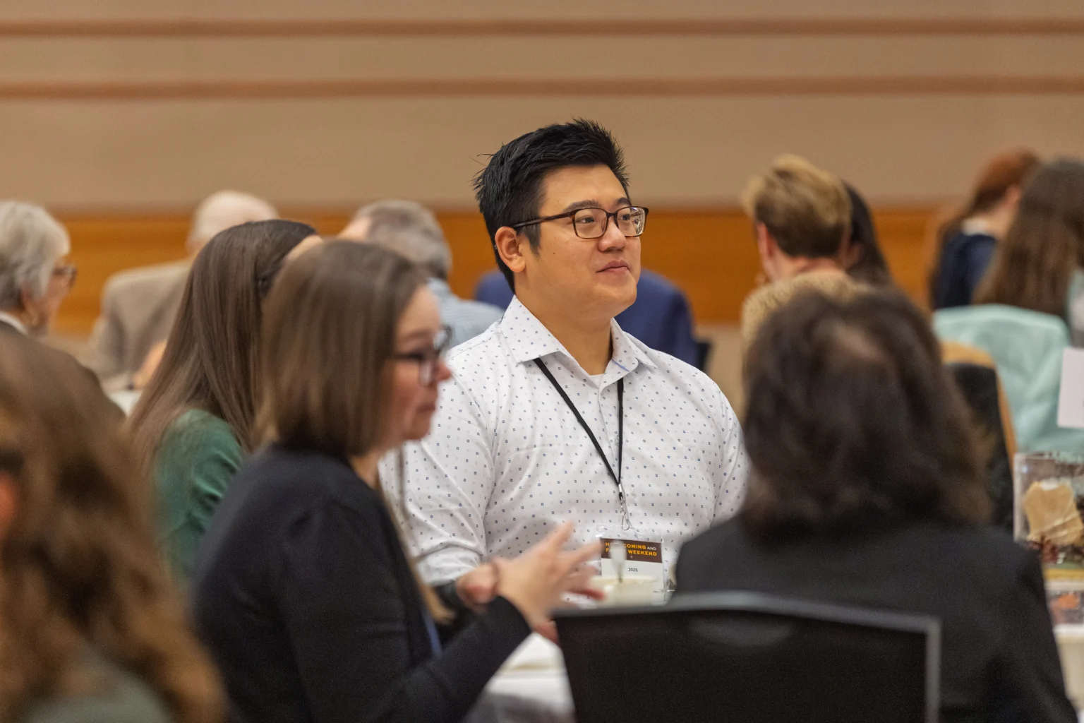 Man in glasses sits at a round table during a conference or meeting, surrounded by other people, including several international students.