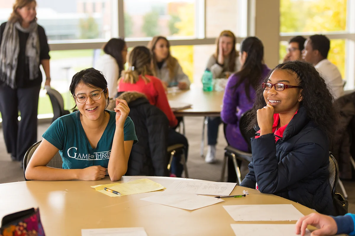 Two international students sit at a table, smiling with papers, while others talk in the bright classroom background.
