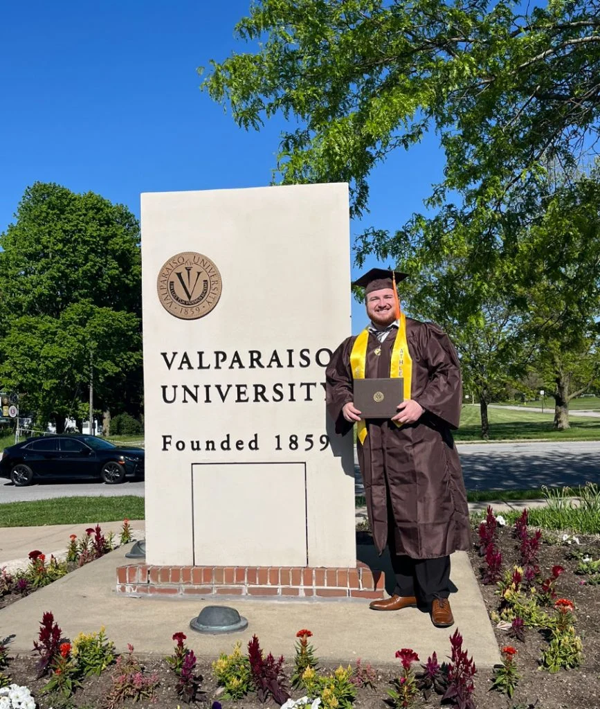 Person in graduation attire holding diploma by Valparaiso University sign, surrounded by flowers and greenery.