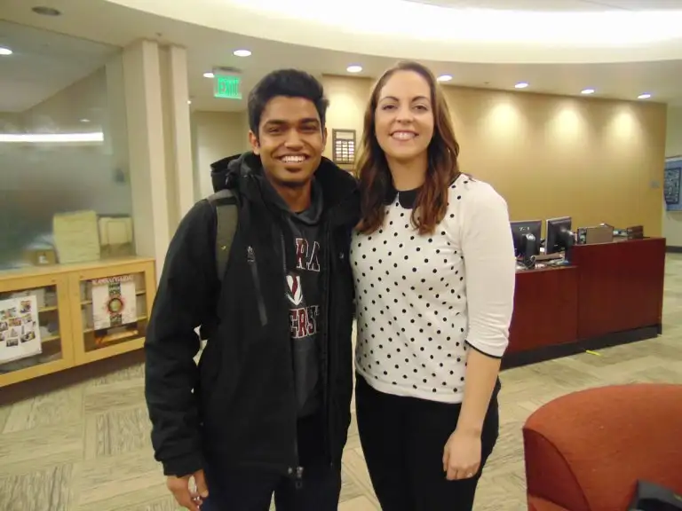Two international students smiling, standing side by side indoors, with office furniture and computers in the background.