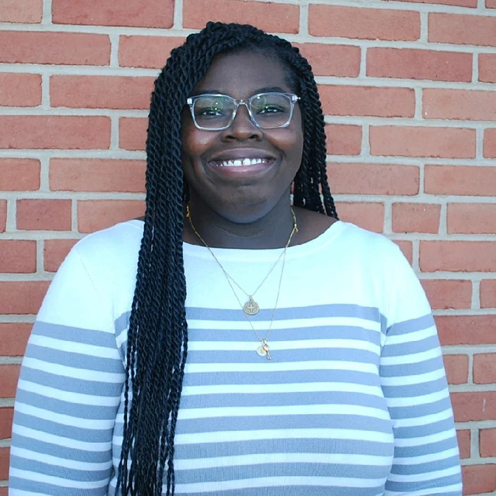 Woman with long braids, glasses, and striped shirt smiles in front of a red brick wall.