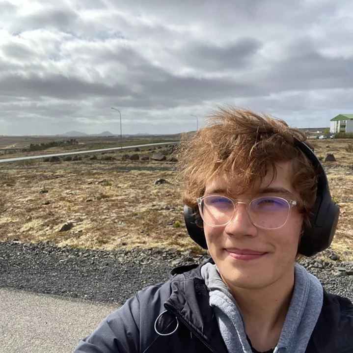 Person wearing glasses and headphones smiles outdoors on a cloudy day near a rural road and rocky field.