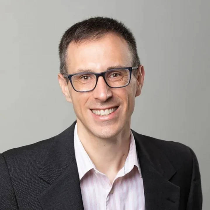 Smiling man in glasses and a dark blazer with a light-colored shirt against a plain background.