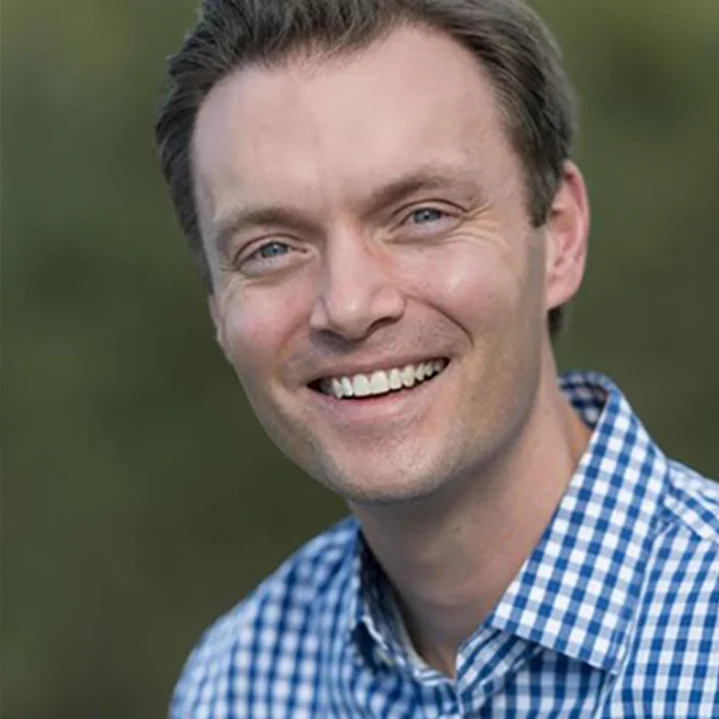 A man smiling outdoors, wearing a blue and white checkered shirt, with a blurred green background.