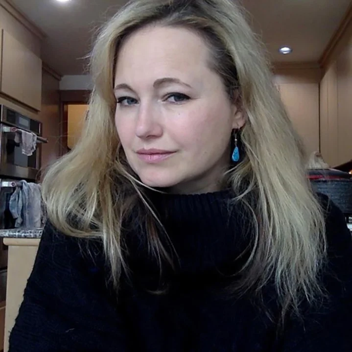 Woman with long blonde hair wearing a black sweater, sitting in a kitchen, looking at the camera.
