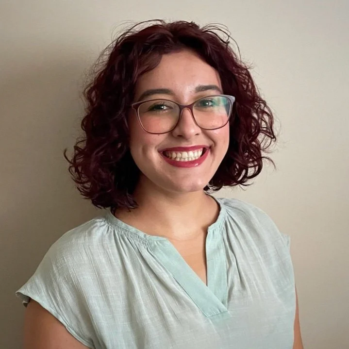 A person with curly dark hair, glasses, and a light blouse smiling in front of a plain background.