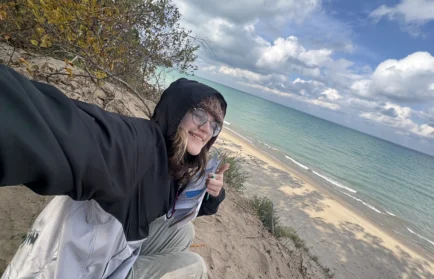 Person in a hooded jacket smiles and gives a thumbs up on a sandy cliff above a beach and blue lake, capturing the spirit of adventure in Nickie Smar’s 2028 photo series, Beacon’s Reflection.