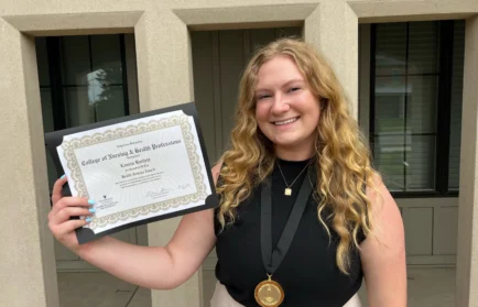 Smiling woman, Lauren Bartlett, holding a certificate and wearing a medal, stands outside a building with beige columns, proudly celebrating her M.S. achievement.