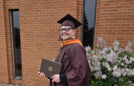 Storm Pendleton, dressed in a maroon graduation gown and cap, holds a diploma while smiling outside near flowers and a brick building—ready to make an impact in healthcare leadership and champion community well-being.