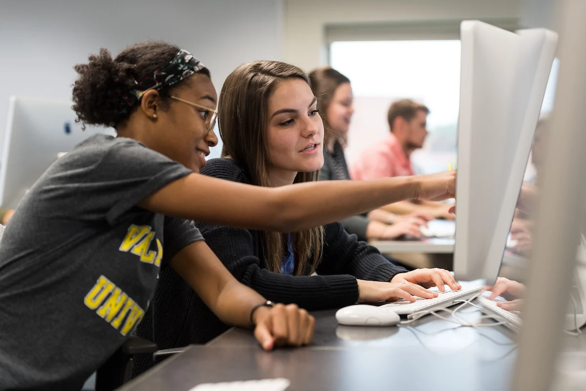Two Valparaiso University students collaborate at a computer in a Communication and Visual Arts classroom, with others working in the background.