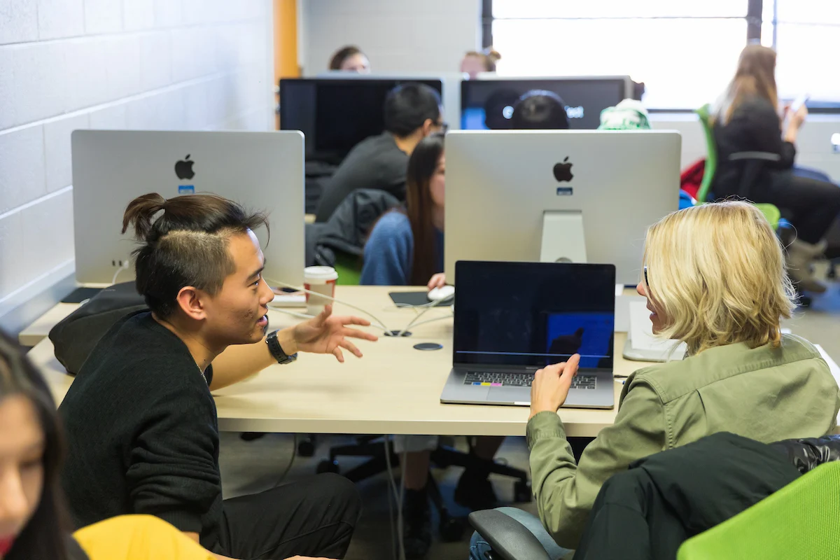 Two people sit at a desk, talking and smiling, showcasing great communication skills, with computers and others working in the background.