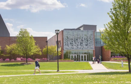 Students walk on a sunny campus lawn in front of a modern building with glass and a patterned facade, where initiatives like AI at Valpo foster Artificial Intelligence Literacy among students.