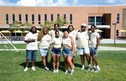 A group of eight people in matching beige shirts and denim shorts stand smiling on grass in front of a brick building, celebrating Valpo BSO and Black History Month.