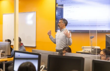 A man teaches a computer class at Valparaiso University, standing in front of monitors and a large screen in a bright classroom focused on the AI Practitioner Certificate.