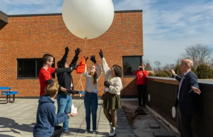 A group of people, including weather profession expert Daniel Murphy, release a large white weather balloon on a sunny rooftop while others watch and take photos.