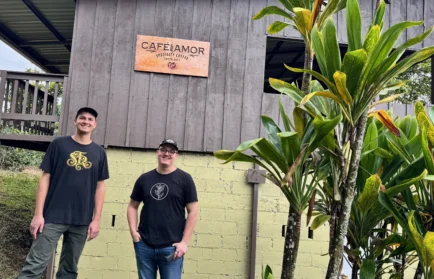 Two people stand smiling in front of a Café Amor sign on a wooden coffee shop, with plants to the right—serving as a welcoming beacon for coffee lovers.