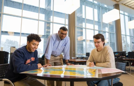 Three students from Valparaiso University look at a large map on a table in a bright, modern room with large windows, exploring concepts from Geospatial Sciences.