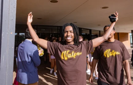 Smiling student in a “Valpo” shirt raises arms outside a building during Opening Convocation, with others from the incoming class in similar shirts nearby.