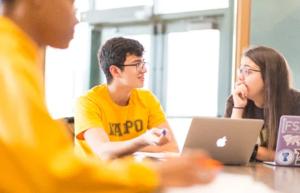 Three students in yellow shirts talk around a table with a laptop in a brightly lit room, sharing insights about what to expect during the first semester of grad school.