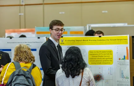 A man in a suit discusses a tornado warning research poster with two women at a science fair, emphasizing the importance of public safety and highlighting recent studies conducted at Valparaiso University.