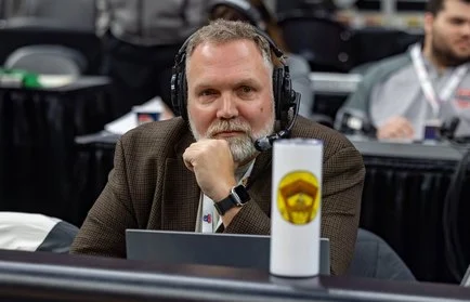 Man with headset and brown jacket sits at table with laptop, resting chin on hand, looking at camera—perhaps deep in thought as Paul Oren prepares for his next Beacon podcast episode.